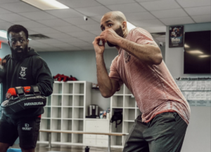 A focused coach Josh in a pink shirt practicing boxing techniques with a coach wearing a black hoodie and red boxing gloves in a martial arts gym. 